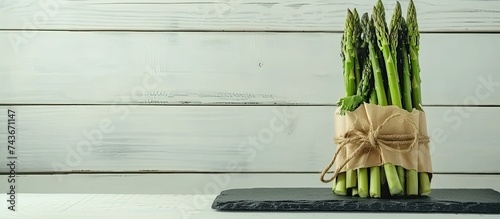 A bunch of fresh green asparagus neatly bundled in brown paper and tied with a rope, placed on a slate dish against a white wooden background.