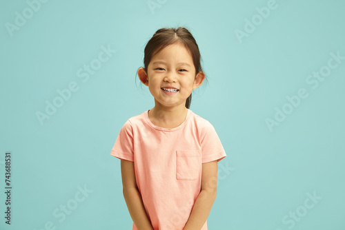 Cheerful portrait of elementary ages Asian girl radiates with a bright smile, expression joyful and happy dressed in a soft peach t-shirt standing against blue isolated background.