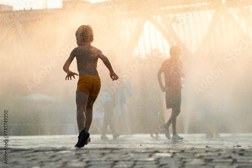 Anonymous silhouette of a child runs through the mist of a city fountain as the sun descends, casting a warm, golden glow over the urban landscape photo