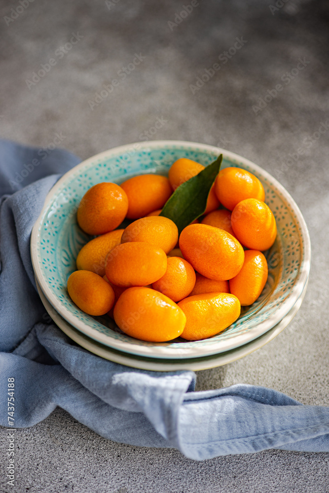Top view of bowl of vibrant, fresh kumquats with a green leaf, placed on a textured surface with a blue napkin Top view of bowl of vibrant, fresh kumquats with a green leaf, placed on a textured surface with a blue napkin