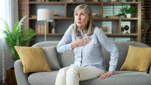 Sick senior gray haired female suffering from heart attack chest tension sitting on sofa in living room at home. Elderly retired woman holding her chest with her hands, needs urgent medical attention photo