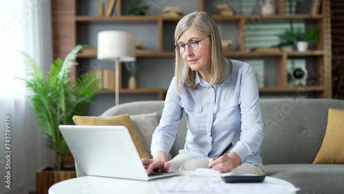 Busy senior gray haired female accountant working doing paperwork sitting in home office. Confident elderly business woman financier makes financial calculations, types on laptop, fills out a form photo
