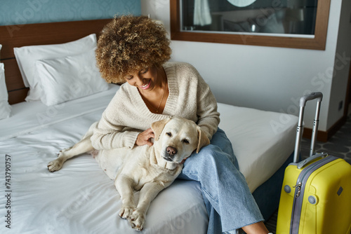 Φωτογραφία young and smiling african american woman cuddling labrador near luggage in pet-f
