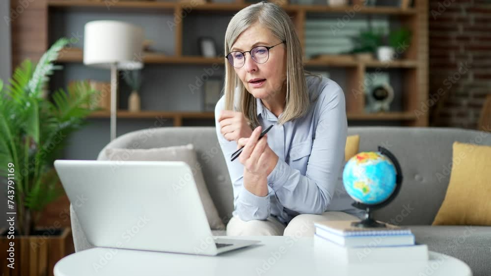 Confident senior gray haired female school teacher in glasses giving video call lesson using laptop sitting in home office. Elderly woman tutor has online training course, talks remotely with students Confident senior gray haired female school teacher in glasses giving video call lesson using laptop sitting in home office. Elderly woman tutor has online training course, talks remotely with students