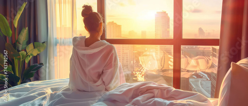 Young woman in bathrobe wake up in modern bedroom open curtains enjoy good morning. Female awake at home or hotel sun shines on her from the big window and city scenery in the window.