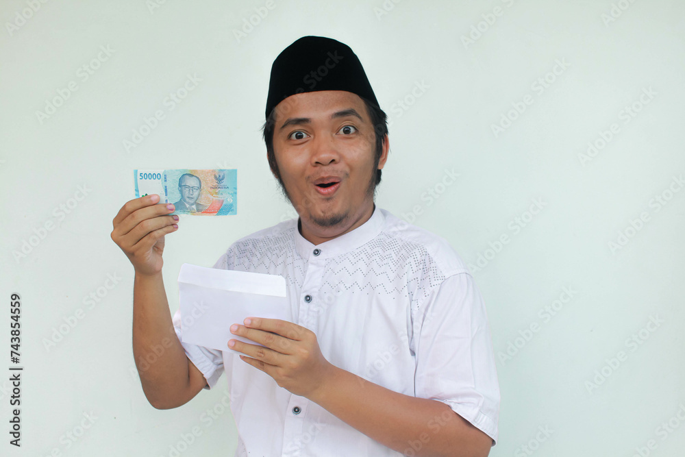A young Asian muslim man holding an envelope and fifty thousand rupiah in cash with an expression of surprise and happiness. THR (Tunjangan Hari Raya) or Religious Holiday Allowance Concept. A young Asian muslim man holding an envelope and fifty thousand rupiah in cash with an expression of surprise and happiness. THR (Tunjangan Hari Raya) or Religious Holiday Allowance Concept.