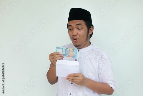 A young Asian muslim man holding an envelope and fifty thousand rupiah in cash with an expression of surprise and happiness. THR (Tunjangan Hari Raya) or Religious Holiday Allowance Concept. photo