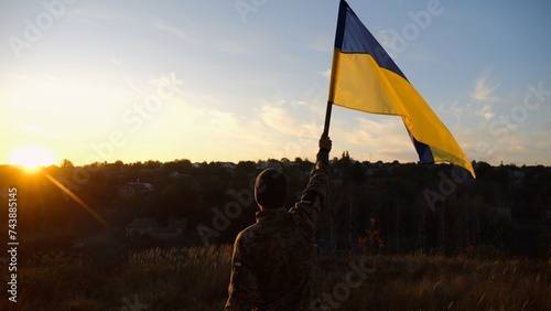 Male soldier stands with raised flag of Ukraine against background of beautiful sunset. Male ukrainian army soldier with a lifted blue-yellow banner in honor of the victory against russian aggression