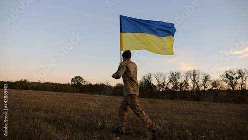 Soldier of ukrainian army running with raised blue-yellow banner on field at dusk. Young male military in uniform jogging with flag of Ukraine at meadow. Victory against russian aggression concept