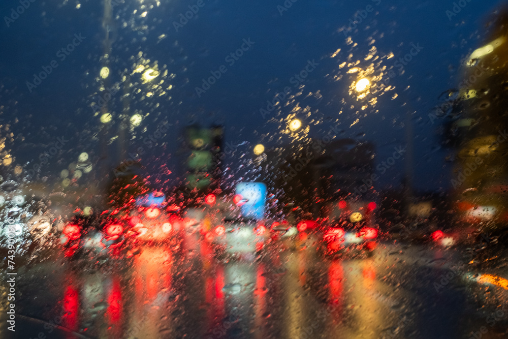 This image captures the blurry lights of a city traffic scene seen through a rain-splattered windshield during nighttime. The droplets on the glass create a soft-focus effect on the red brake lights
