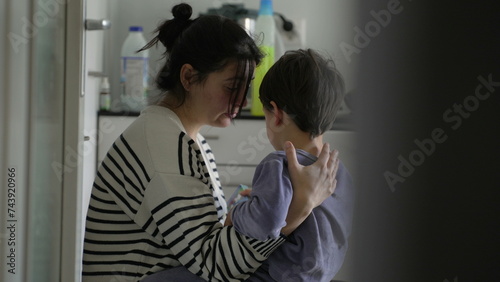 Caring mother consoling crying little son in candid kitchen scene. Mom embracing hurt child, candid and tender photo