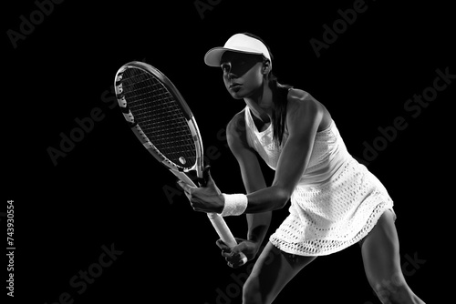 Focused athlete in tennis attire awaiting opponent's serve against black studio background. Monochrome filter. Concept of women in sport, active lifestyles, tournaments and events, energy, movement. © Lustre