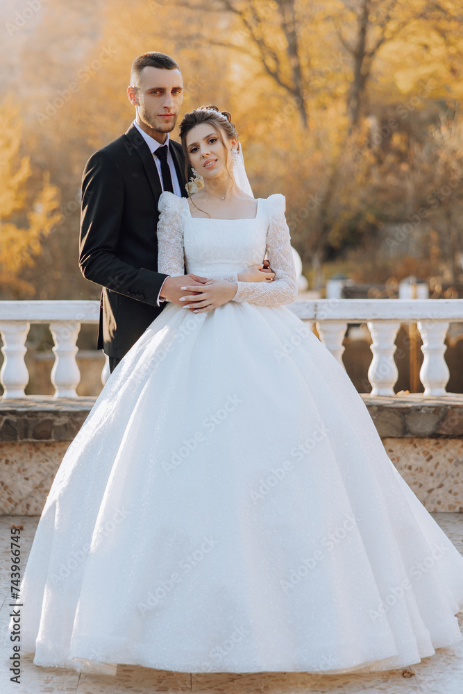 Stylish, young groom and beautiful bride in a long white dress and a long veil with a bouquet in their hands, hugging in the park in the autumn nature. Wedding portrait of newlyweds.