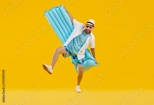 Funny overjoyed excited man wearing beach clothes with open mouth holding inflatable mattress isolated on a studio yellow background. Happy tourist is going on summer holiday trip. Vacation concept.