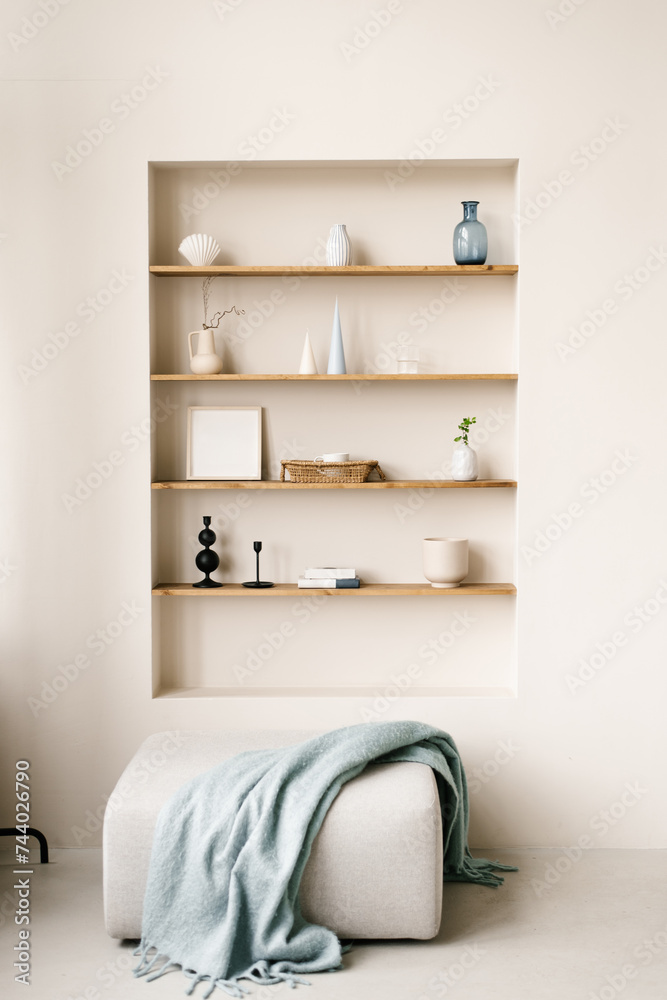 Modern living room with pouf with blue blanket. Built-in shelves in the wall with decor, personal accessories, ceramics in Scandinavian minimalist style