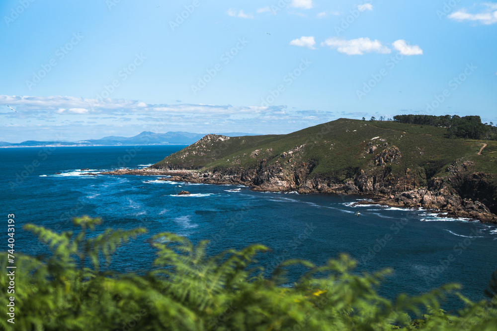 Captivating Ons Island scene: lush green ferns in the foreground, azure sea, rocky cliffs, Pontevedra estuary, and a summer sky with scattered clouds from the Buraco do Inferno path.