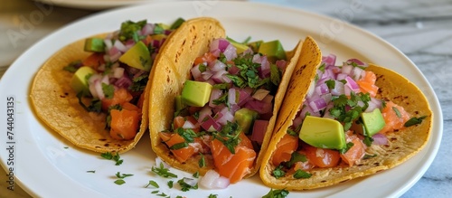 A photo featuring three homemade tacos made with salmon, onions, cucumbers, and avocado, arranged neatly on a white plate and topped with fresh vegetables.