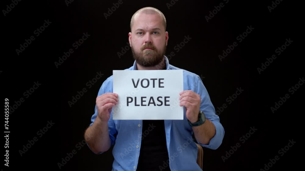 General view bald bearded man in blue shirt look in camera and shows piece of paper with the inscription VOTE PLEASE. Call for voting in elections.