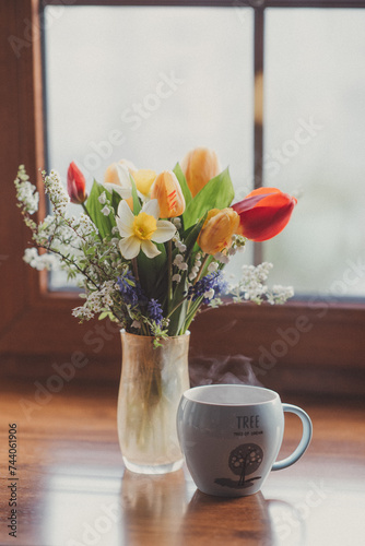a spring bouquet of tulips  daffodils  spirea stands on the windowsill with a mug of coffee. the spring atmosphere of the house. retro postcard. the aesthetics of gardening
