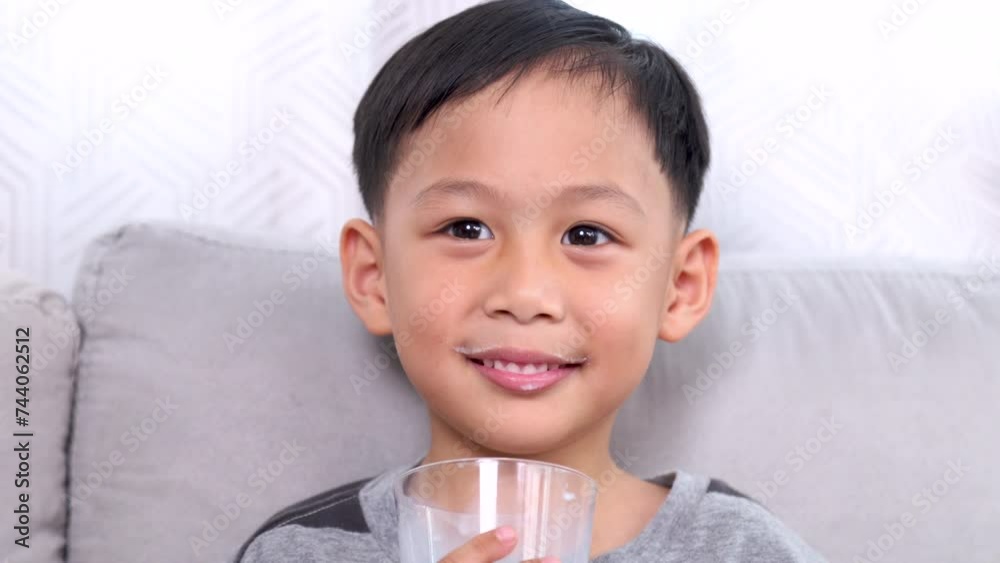 Close-up adorable Asian preschool boy drinks dairy milk from glass, smiling kid happy with protein beverage enjoy sitting in living room on weekend playing alone laughing watching game talking to mom Close-up adorable Asian preschool boy drinks dairy milk from glass, smiling kid happy with protein beverage enjoy sitting in living room on weekend playing alone laughing watching game talking to mom