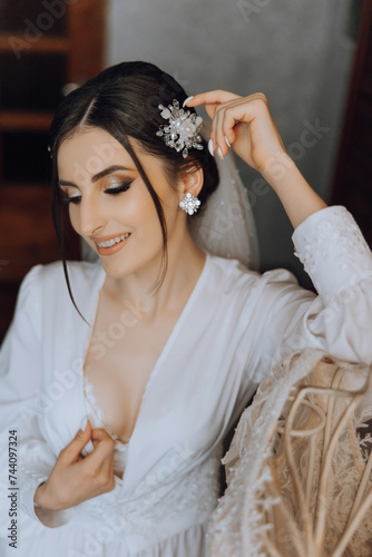 an incredibly beautiful bride in the morning in a white robe and a long veil sits on the bed in her bedroom. The bride poses in the morning before the wedding ceremony.