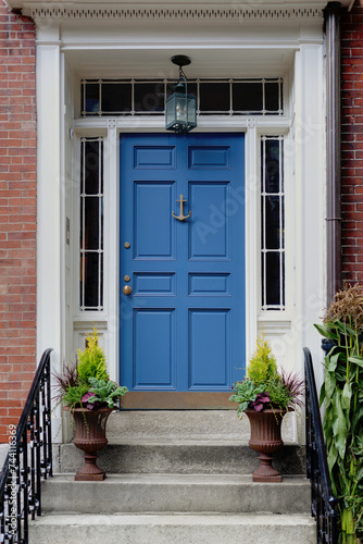 A vibrant blue front door features sidelight windows on each side and a transom window above. Two potted plants outside the door in New England. photo