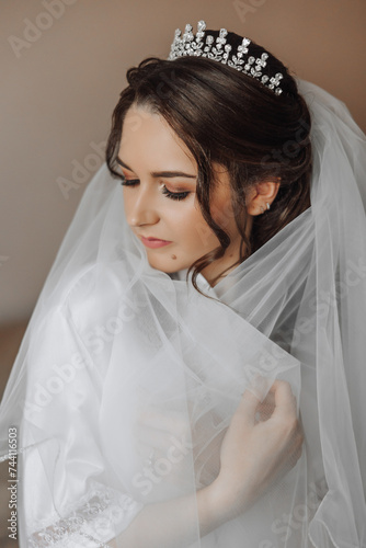 portrait of an incredibly beautiful girl bride in a white robe in the bedroom, the bride poses holding her veil in her hands and covers herself with it.