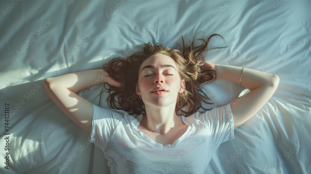 Fototapeta premium Top view of a relaxed young woman lying on bed with hands behind head, basking in morning sunlight.