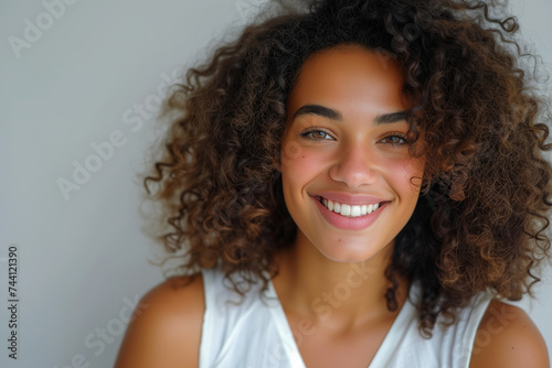 Radiant multiethnic woman with curly hair and a beaming smile  wearing a white tank top on a light background.