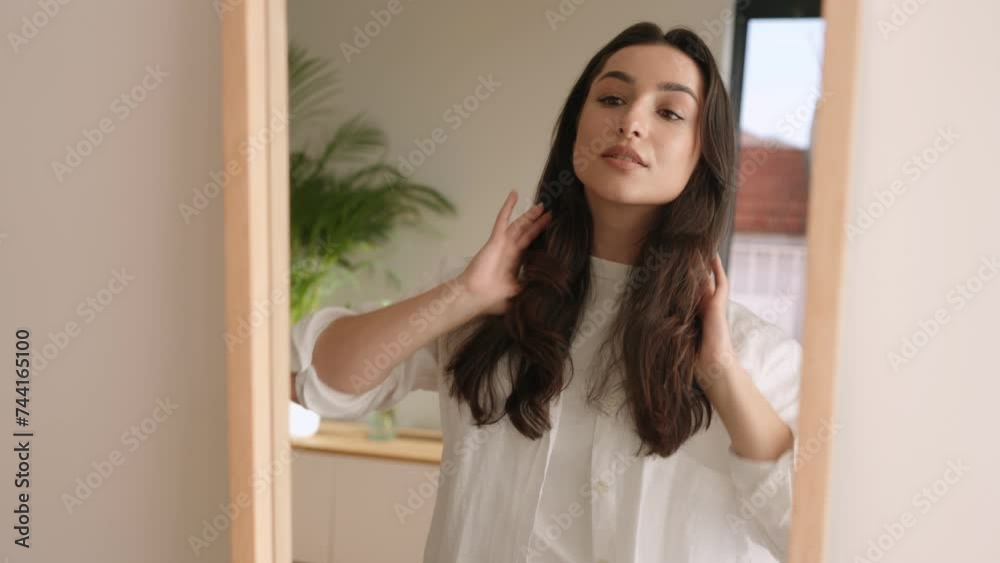 A charming lady with a content expression looks into a mirror, touching her hair gently, in a well-lit interior space. A charming lady with a content expression looks into a mirror, touching her hair gently, in a well-lit interior space.