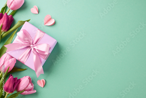 Tokens of gratitude: curated surprises for her. Top view shot of gift box with pink satin ribbon, pink paper hearts, tulips on teal background with space for special occasion greetings messages photo