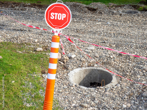 A stop sign in a park in front of an open manhole. Danger lies ahead. A lopsided sign. There is no passage.