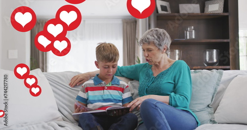 Image of heart icons over caucasian grandmother and grandson reading book in livin groom