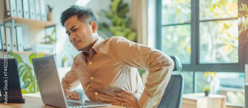 Asian businessman having sick back pain after extra time working with laptop in office. AI generated photo