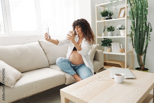 Pregnant woman blogger with a jar of pills and vitamins sits on the couch at home and takes pictures of herself on the phone, maintaining pregnancy and replenishing vitamins and minerals