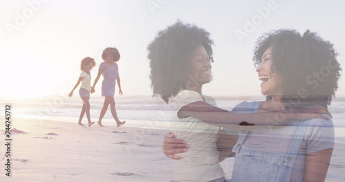 Composite of happy african american mother and daughter embracing, and raising arms walking on beach
