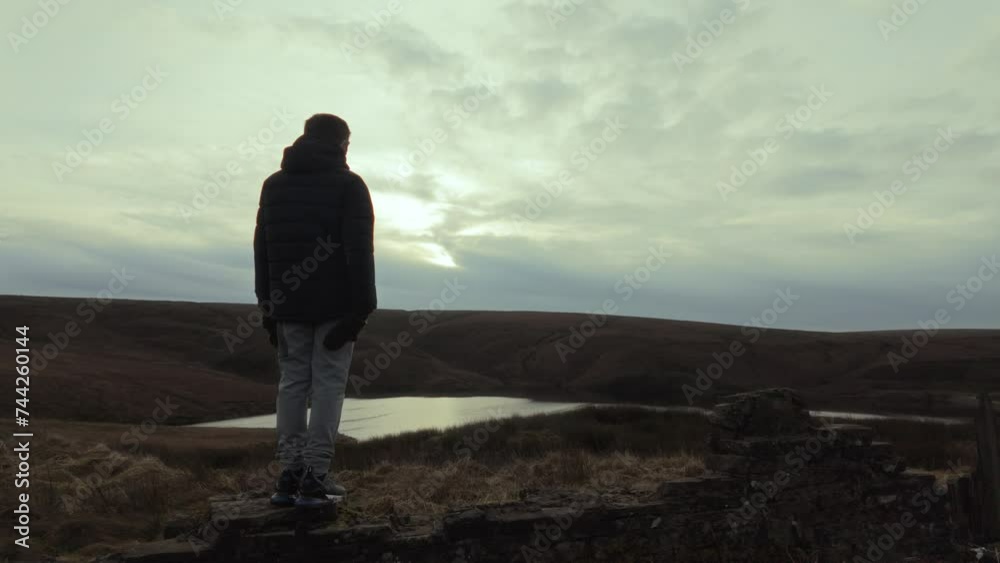 Young boy standing and looking out on to the winter landscape, with moorlands, fens, a lake and stormy winters sky. Sunset view of the Yorkshire landscape