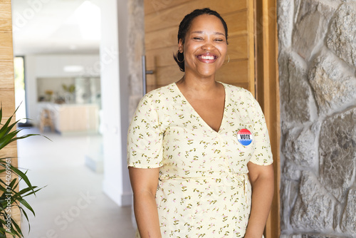 A smiling young biracial woman stands in a bright hallway, wearing a vote badge, with copy space