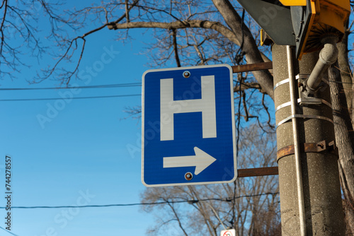 projecting road sign with capital H and arrow pointing left on a blue sky with bare trees in winter