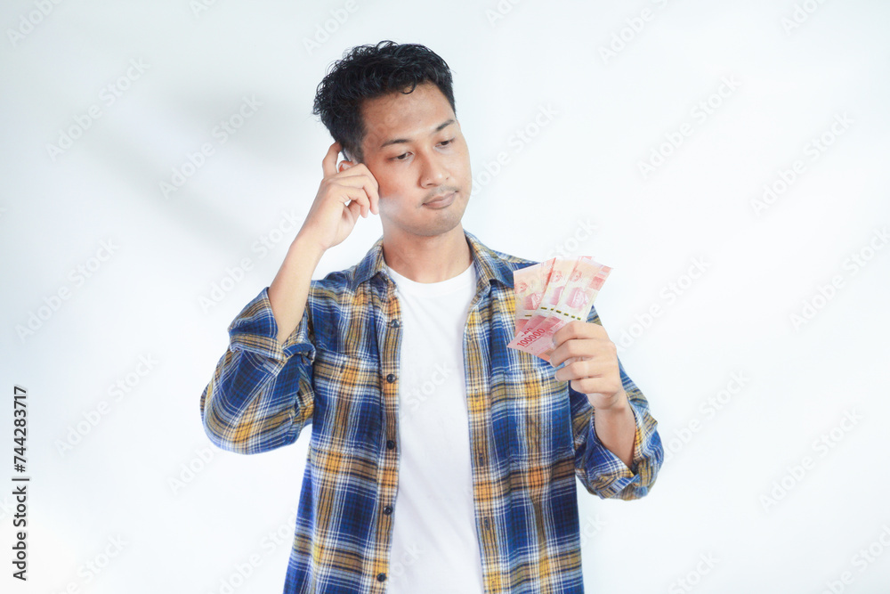 Pensive young Asian student man in casual clothes with backpack Holding fan of cash money in rupiah banknotes isolated on white background. High school university college concept Pensive young Asian student man in casual clothes with backpack Holding fan of cash money in rupiah banknotes isolated on white background. High school university college concept