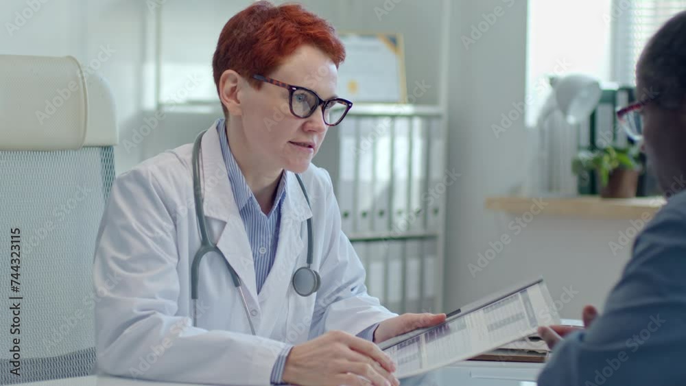 Redhead female doctor checking medical records and speaking with male patient about his disease symptoms during appointment in clinic