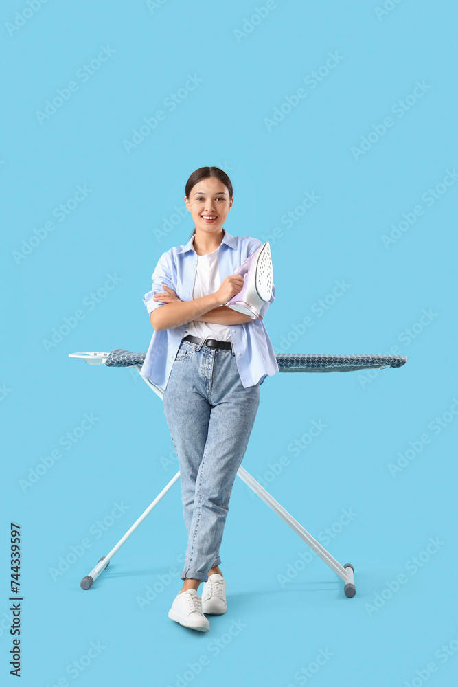 Happy Asian woman with iron standing near ironing board on blue background