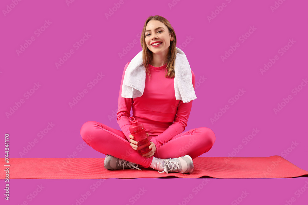 Beautiful young happy woman in sportswear with bottle of water and yoga mat on purple background Beautiful young happy woman in sportswear with bottle of water and yoga mat on purple background