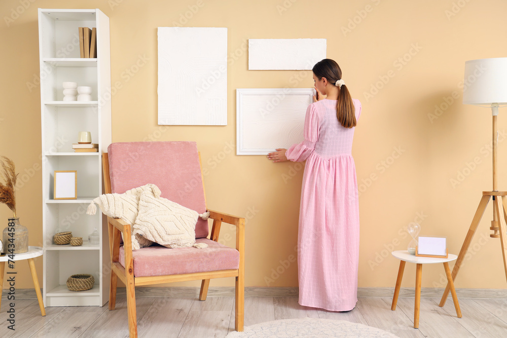 Young woman hanging stylish painting on beige wall in living room, back view Young woman hanging stylish painting on beige wall in living room, back view