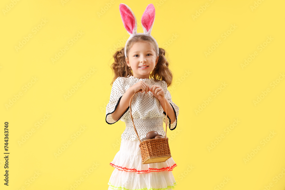 Cute little girl in bunny ears with basket of chocolate Easter eggs on yellow background