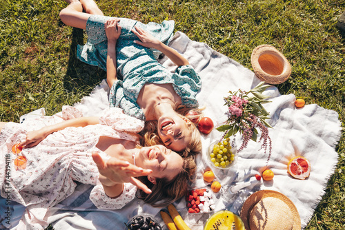 Two young beautiful smiling hipster female in summer sundress and hats. Carefree women making picnic outside. Positive models sitting on plaid on grass, drinking champagne, eating fruits. Top view