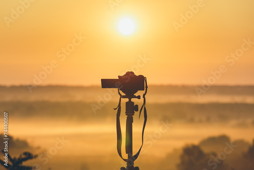 A professional photographer's camera on a tripod looks at the sunrise. Countryside landscape with camera on tripod, sunrise and morning fog. photo