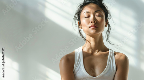 Free Download Beautiful and stylish young Asian woman in white tank top with her eyes closed, meditate and contemplate in white room in sunlight from window - GFXLoad