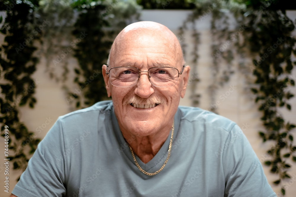 Close-up Portrait of Happy 70 Years Old Senior Man Smiling Looking at Camera. Relaxed Grandfather in Casual Clothing and Eyeglasses Sitting Inside Close-up Portrait of Happy 70 Years Old Senior Man Smiling Looking at Camera. Relaxed Grandfather in Casual Clothing and Eyeglasses Sitting Inside