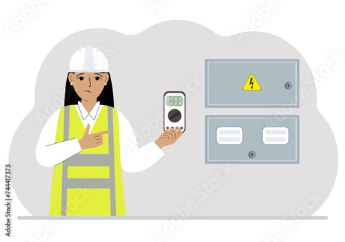 A female electrical engineer holds a digital multimeter in his hand and stands next to an electrical cabinet.